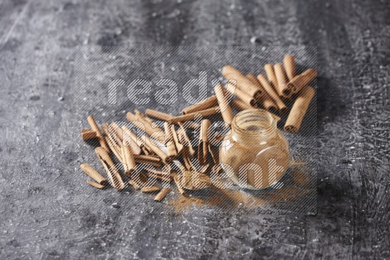 Herbal glass jar and a metal spoon full of cinnamon powder surrounded by cinnamon sticks on textured black background