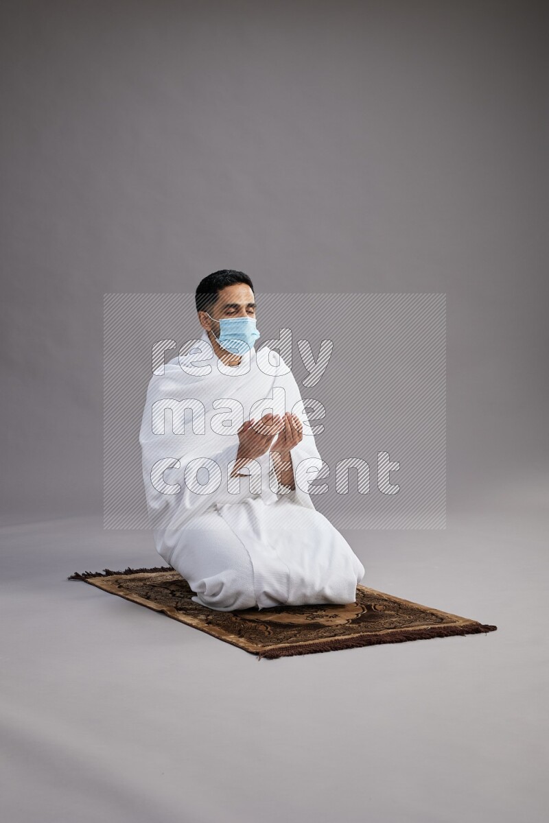 A man wearing Ehram with face mask sitting on floor performing dua'a on gray background