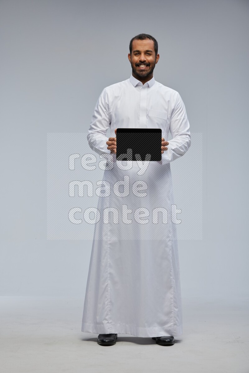 Saudi man wearing Thob standing showing tablet to camera on Gray background