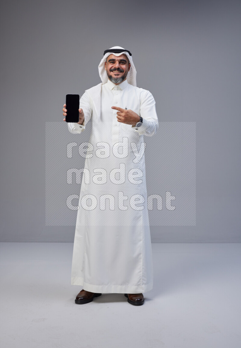 Saudi man Wearing Thob and white Shomag standing showing phone to camera on Gray background