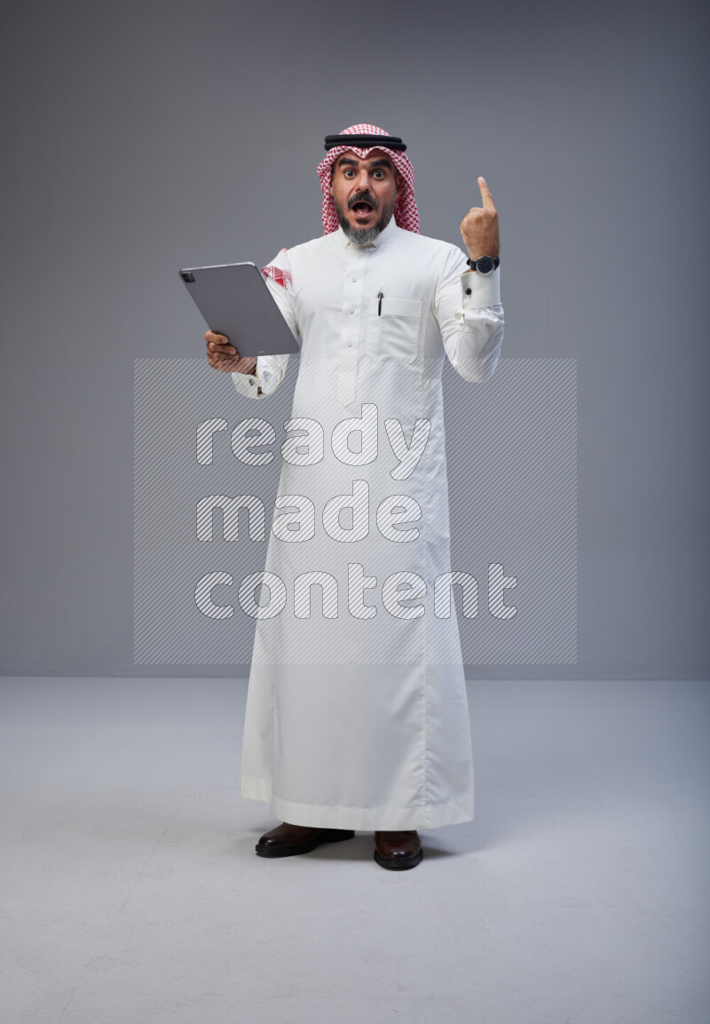 Saudi man Wearing Thob and red Shomag standing working on tablet on Gray background