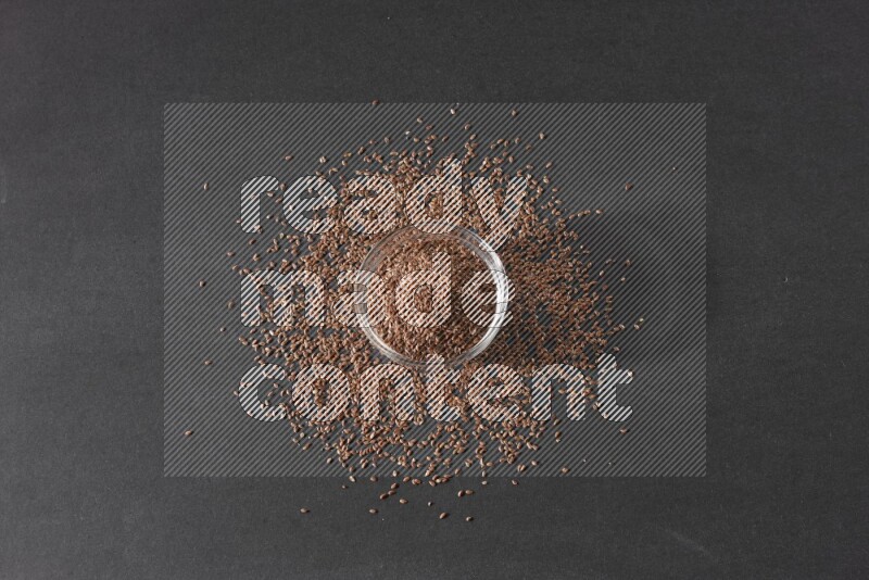 A glass bowl full of flaxseeds surrounded by the seeds on a black flooring
