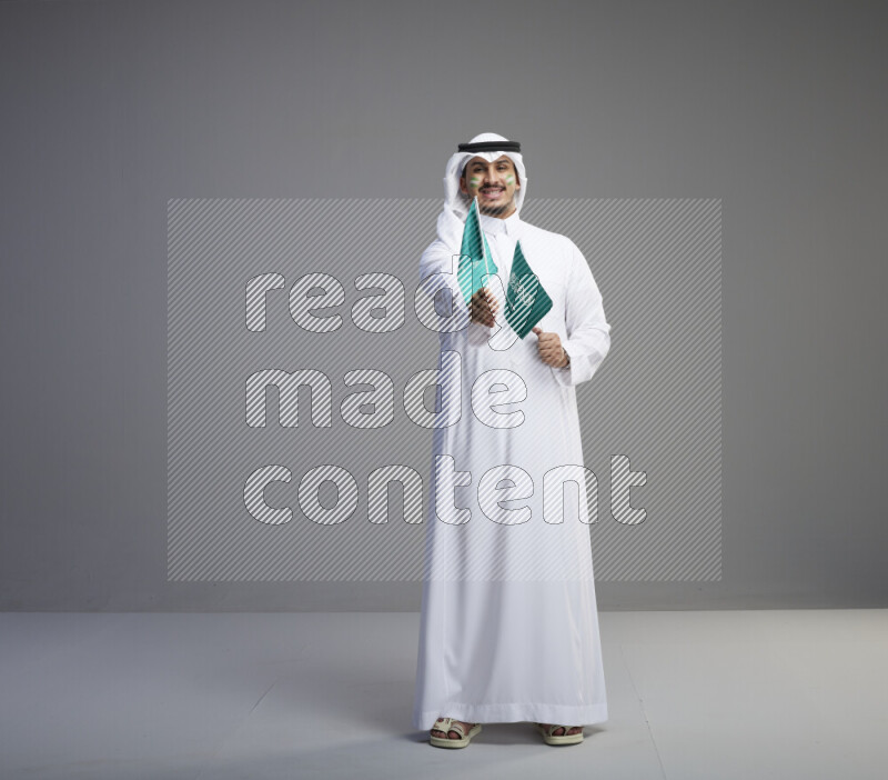 A Saudi man standing wearing thob and white shomag with face painting raising small saudi flag on gray background