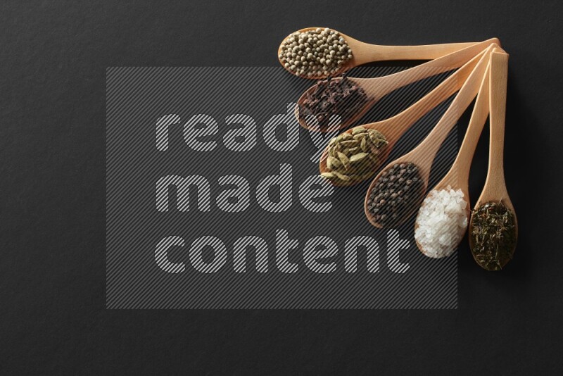 wooden spoons filled with white peppers, cloves, cardamom, salt, black peppers and basil on black flooring