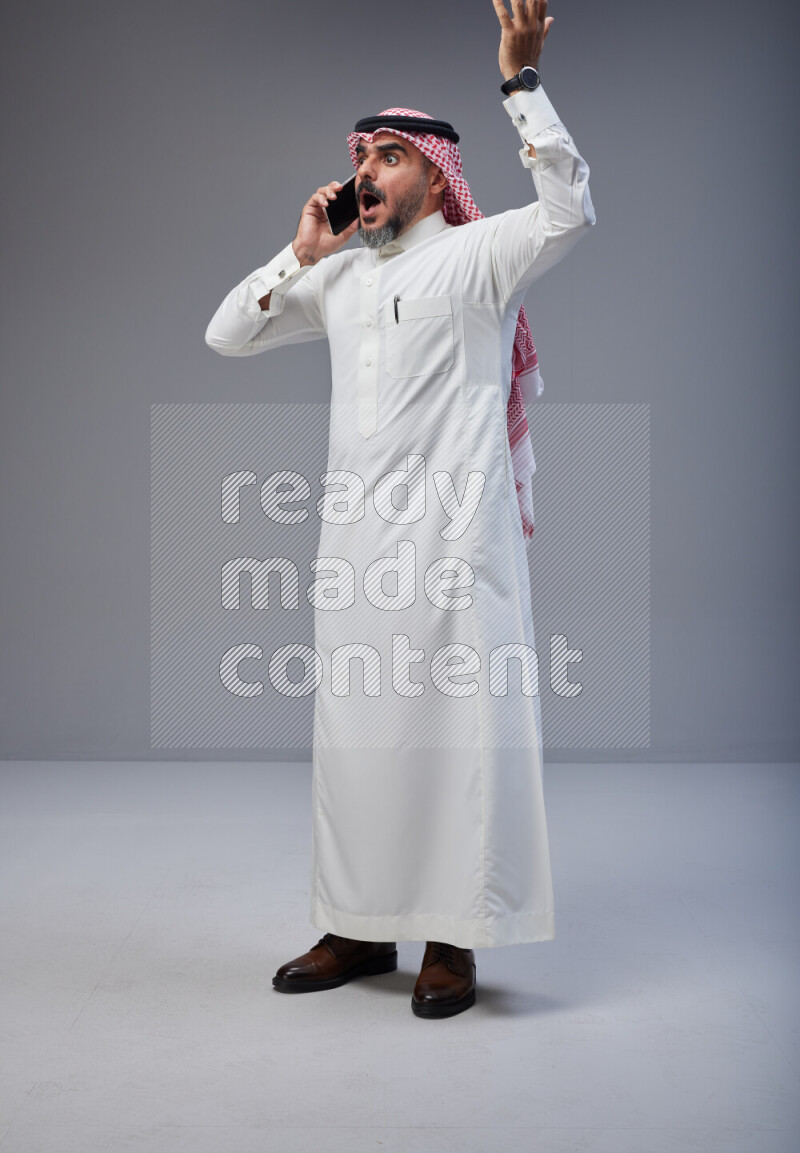 Saudi man Wearing Thob and red Shomag standing talking on phone on Gray background