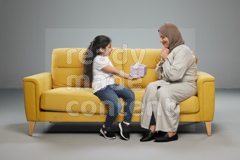 A girl sitting on a yellow sofa and giving a gift to her mother on gray background