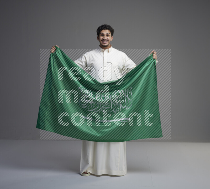 A Saudi man standing wearing thob holding big Saudi flag on gray background