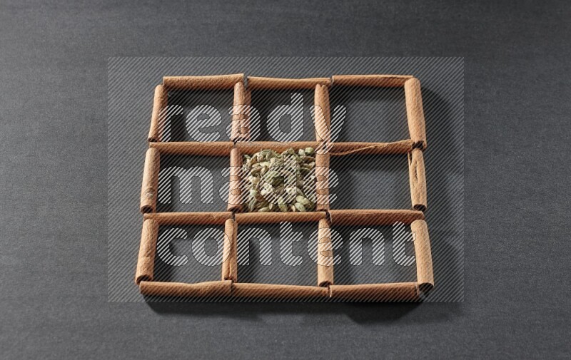 9 squares of cinnamon sticks full of cardamom in the middle surrounded by nutmeg, cinnamon, bay laurel leaves, cloves, cumin, dried ginger, dried basil and star anise on black flooring