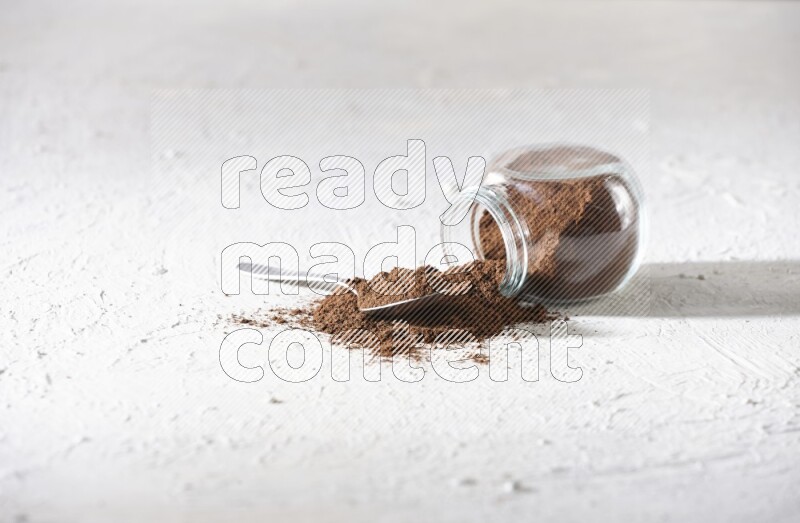 A flipped glass spice jar and a metal spoon full of cloves powder and powder came out of the jar on textured white flooring