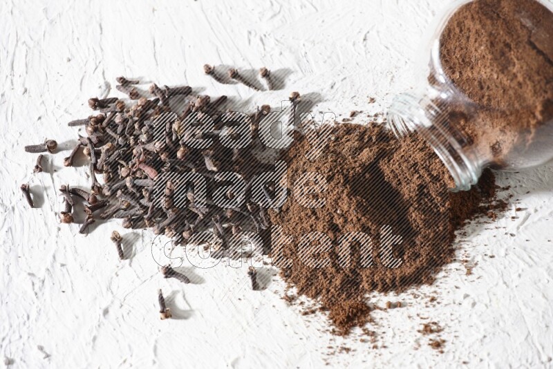 A glass spice jar full of cloves powder flipped and powder came out of it with cloves spread on textured white flooring