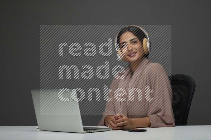 A Saudi woman Sitting on her desk Typing on her laptop wearing a headset  on a Gray Background wearing Brown Abaya