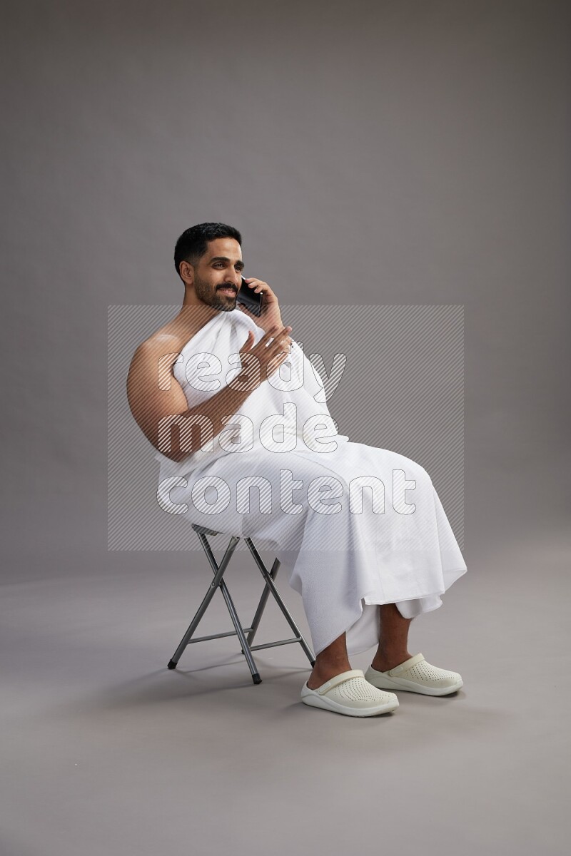 A man wearing Ehram sitting on chair talking on the phone on gray background