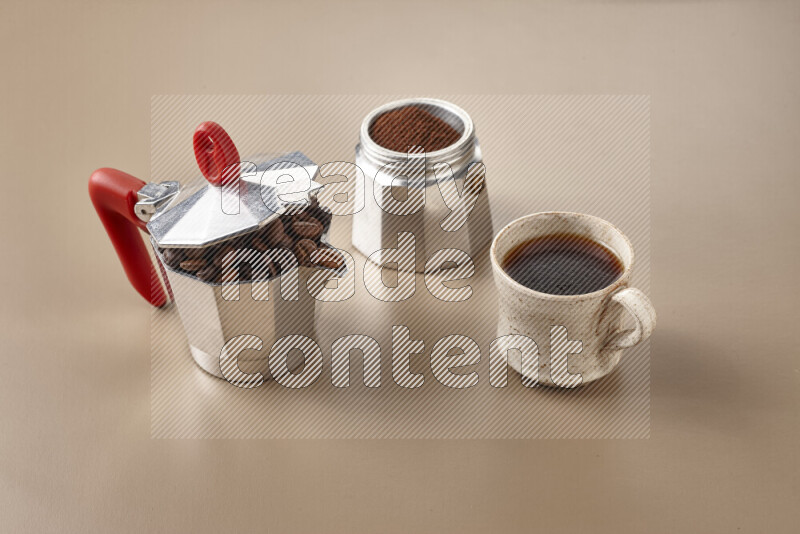 A moka pot with red handle surrounded by roasted coffee beans on beige background