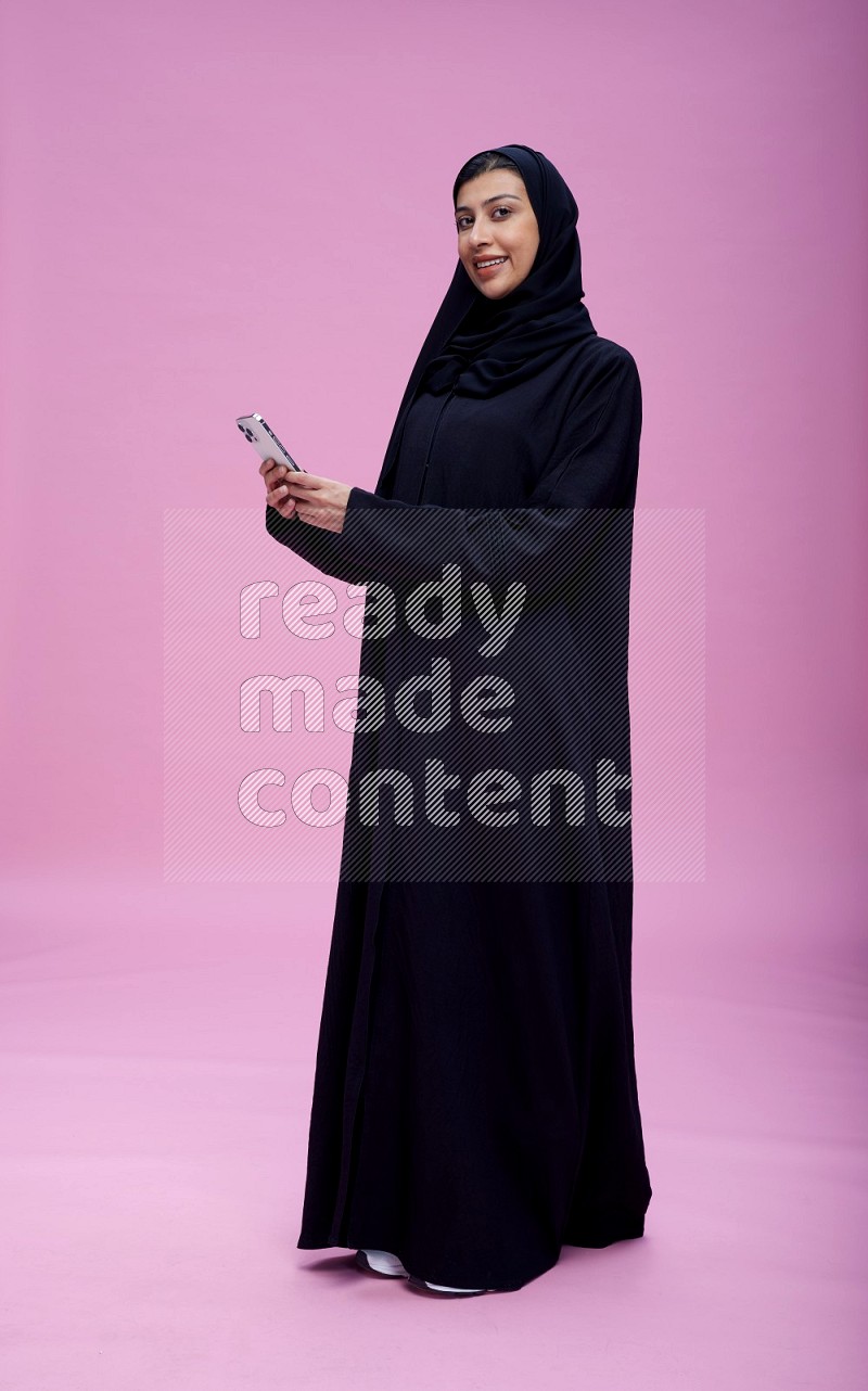 Saudi woman wearing Abaya standing texting on phone on pink background