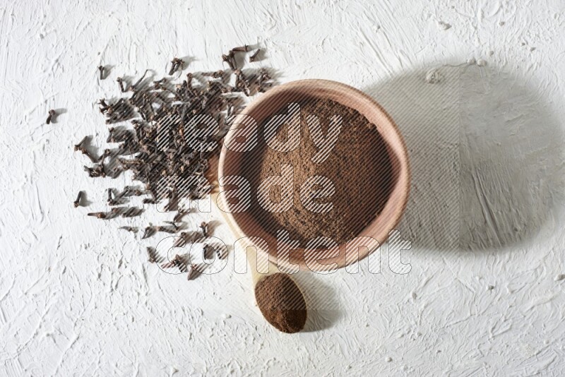 A wooden bowl and wooden spoon full of cloves powder with cloves spread on textured white flooring