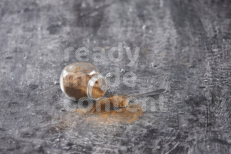 Herbal glass jar full of cinnamon powder flipped and a metal spoon on textured black background