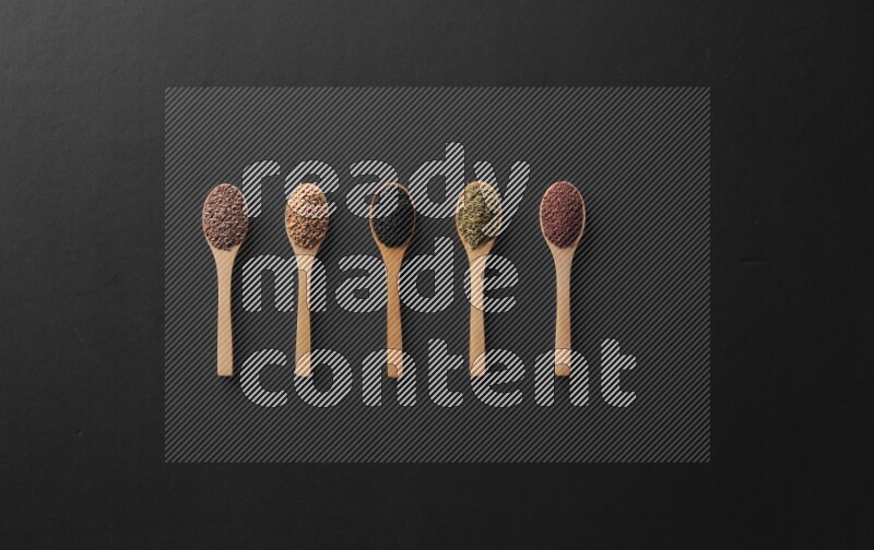 wooden spoons filled with white peppers, mustard seeds, black seeds, garden cress, cumin and flax on black flooring