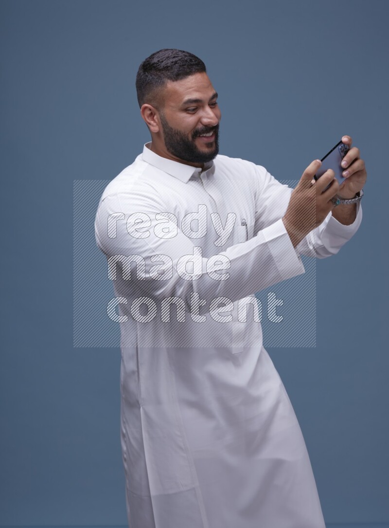 A man Playing a Game on his Smartphone  on Blue Background wearing Saudi Thob