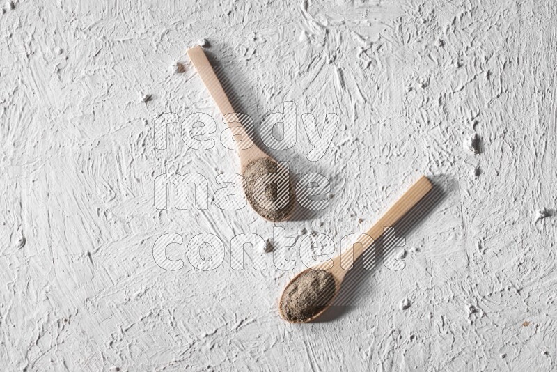 2 wooden spoons full of black pepper powder on a textured white background