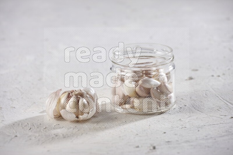 A glass jar full of garlic cloves with a whole garlic bulb beside it on a textured white flooring