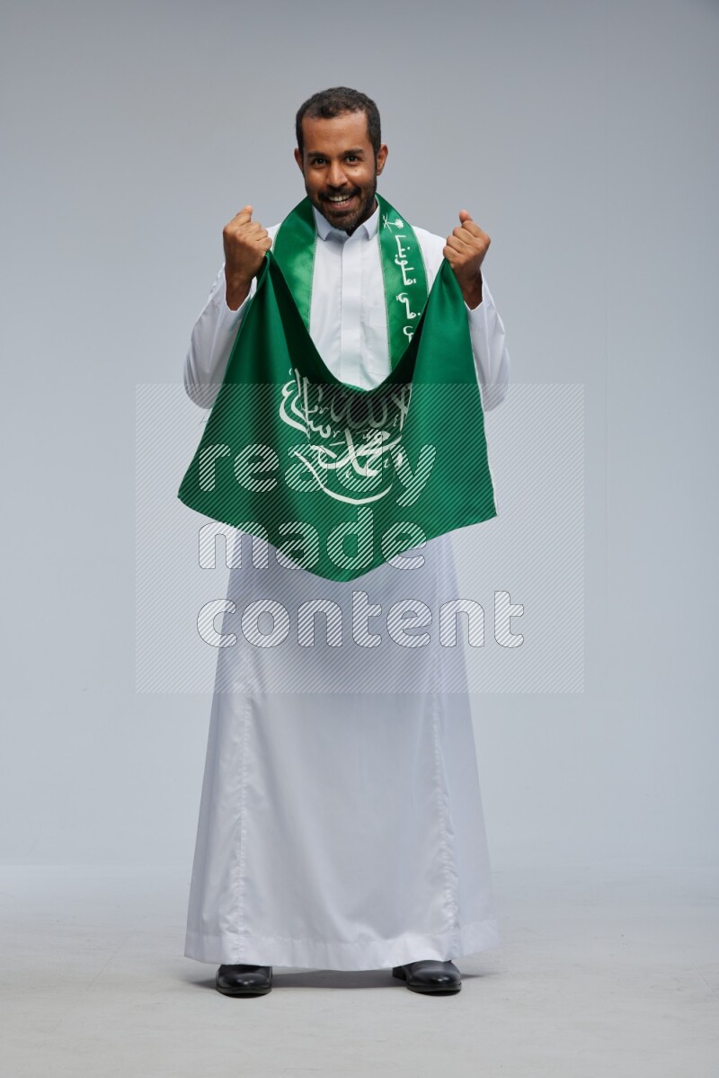 Saudi man wearing Thob standing holding Saudi flag on Gray background