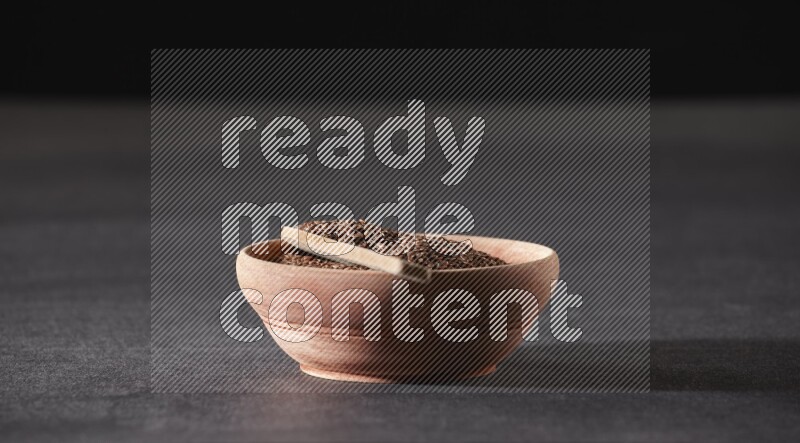 A wooden bowl full of flaxseeds with wooden spoon full of the seeds on it on a black flooring