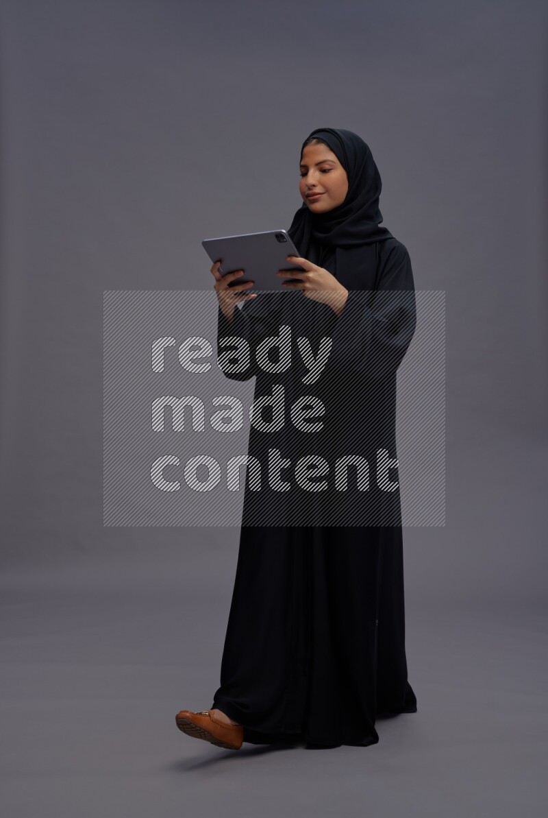Saudi woman wearing Abaya standing working on tablet on gray background