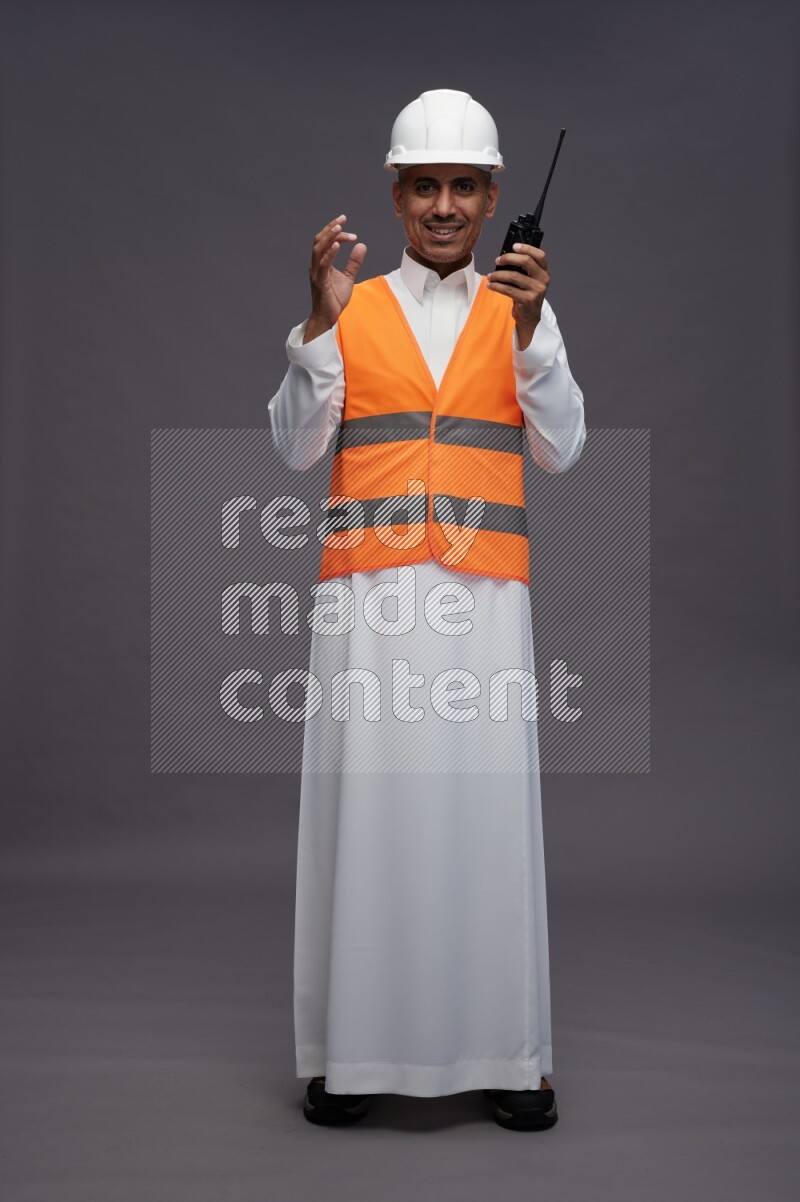 Saudi man wearing thob with engineer vest standing holding walkie-talkie on gray background