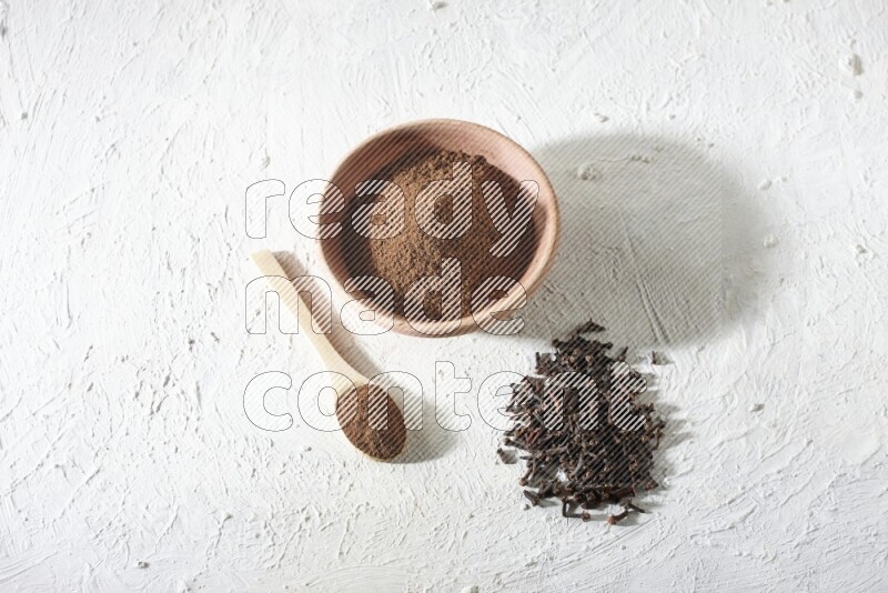A wooden bowl and wooden spoon full of cloves powder with cloves spread on textured white flooring