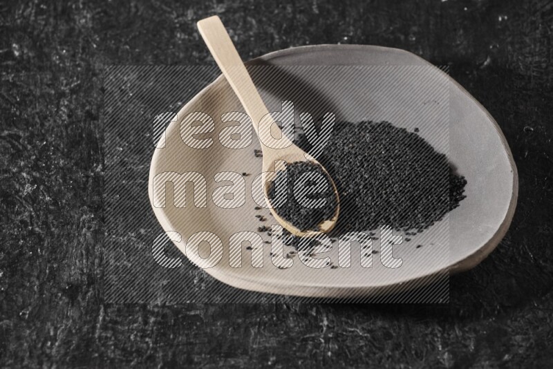A multicolored pottery plate full of black seeds and wooden spoon full of seeds on a textured black flooring
