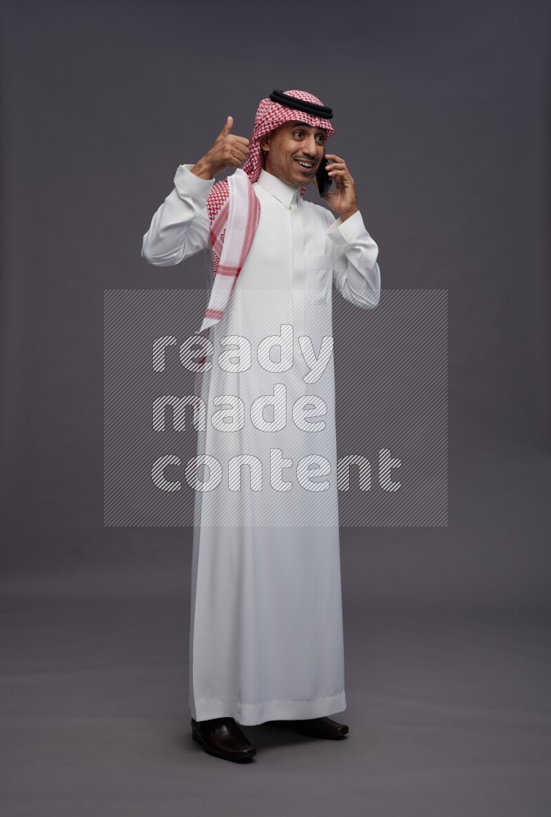 Saudi man wearing thob and shomag standing talking on phone on gray background