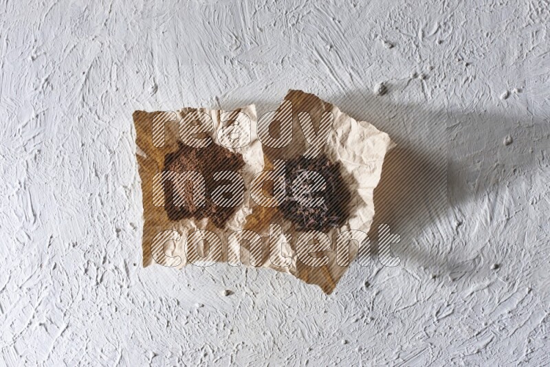 Cloves powder and cloves on 2 crumpled pieces of paper on a textured white flooring