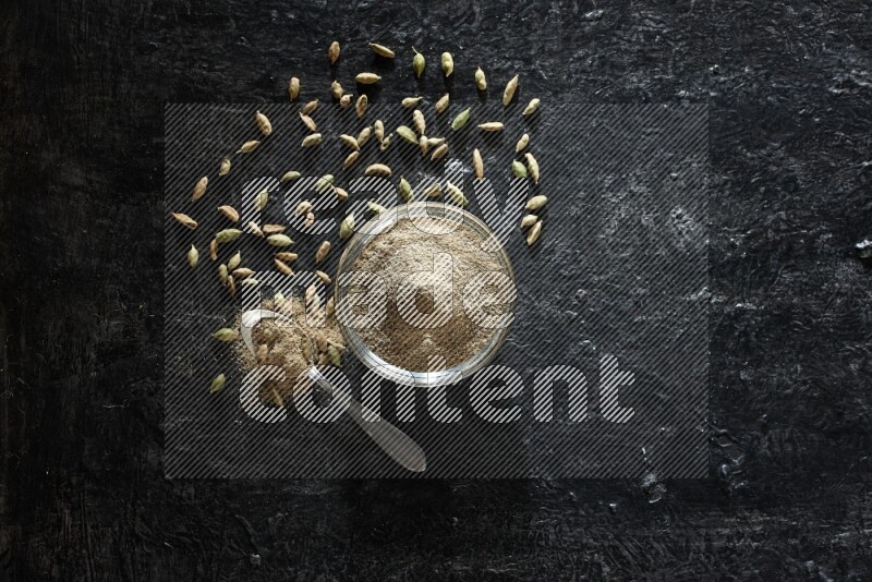 A glass bowl and a metal spoon full of cardamom powder with cardamom seeds beside them on textured black flooring
