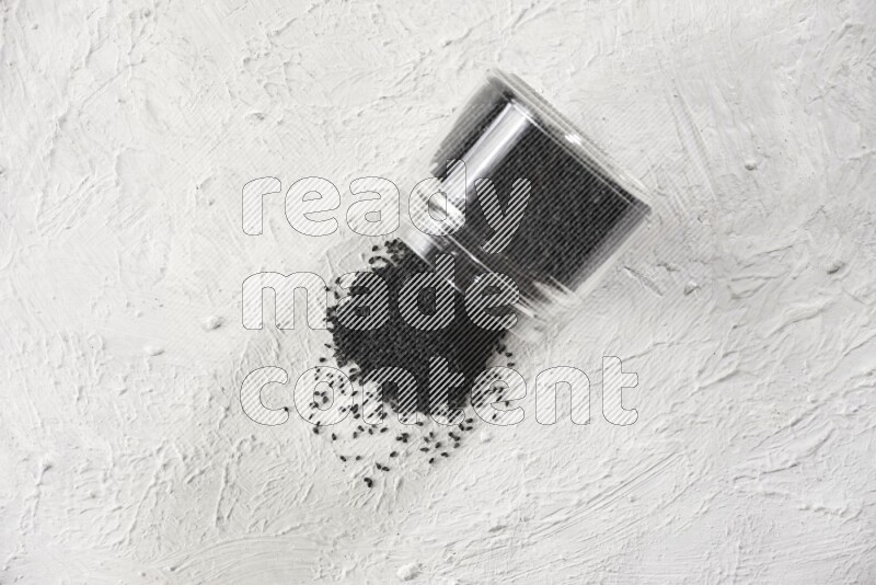 A glass jar full of black seeds and the jar flipped and seeds spread on a textured white flooring