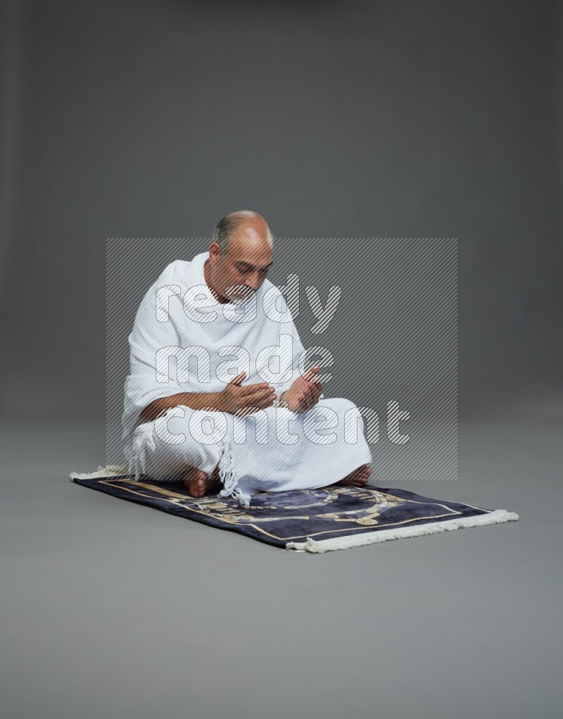 A man wearing Ehram sitting on prayer mat dua'a on gray background