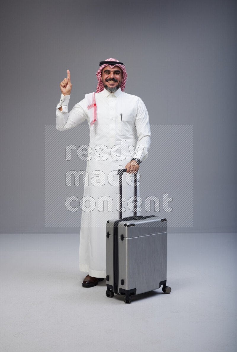 Saudi man wearing Thob and red Shomag standing holding Travel bag on Gray background