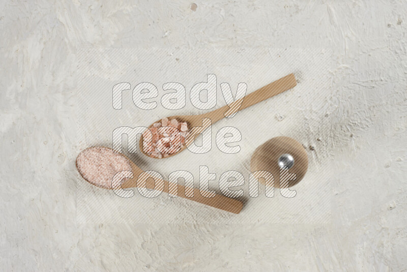 2 wooden spoons filled with fine and coarse salt with wooden grinder beside them on white background