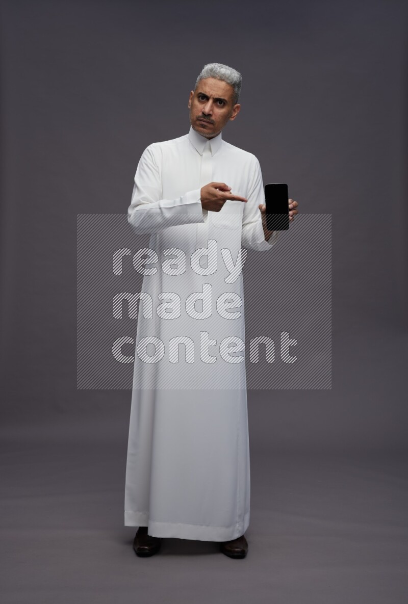 Saudi man wearing thob standing showing phone to camera on gray background