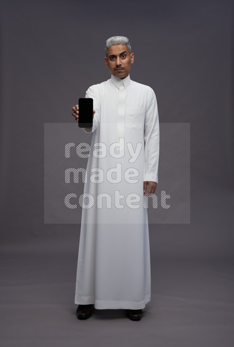 Saudi man wearing thob standing showing phone to camera on gray background