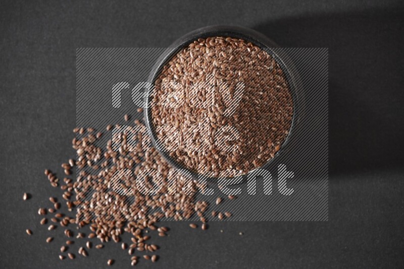 A black pottery bowl full of flaxseeds surrounded by the seeds on a black flooring