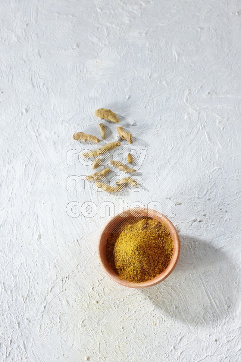 A wooden bowl full of turmeric powder with dried whole fingers on textured white flooring