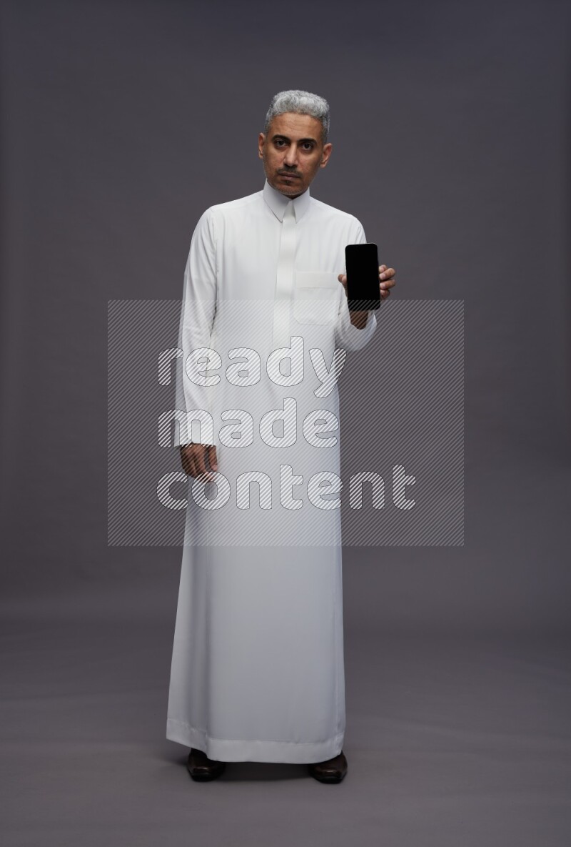 Saudi man wearing thob standing showing phone to camera on gray background