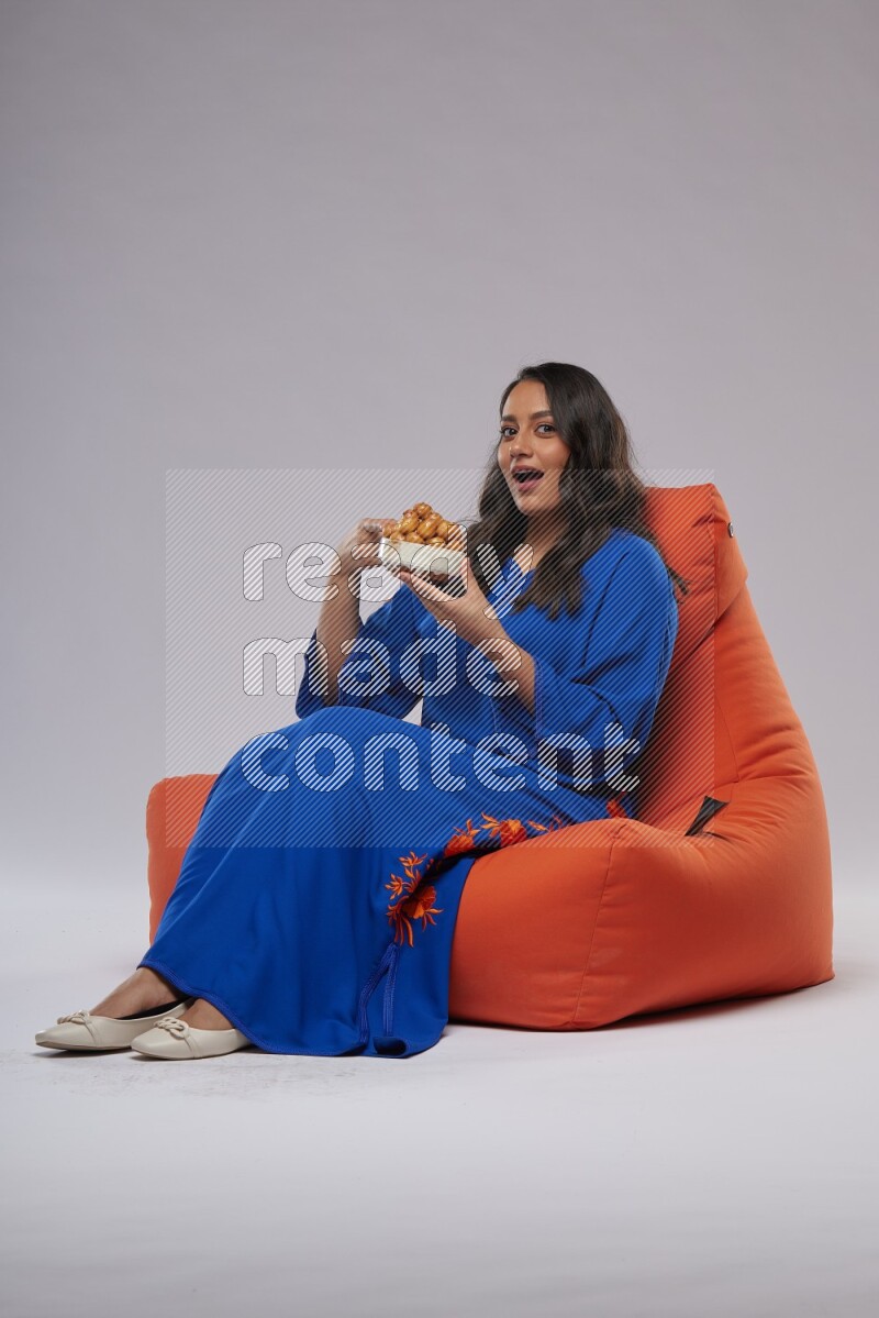 A Woman sitting on an orange beanbag wearing Jalabeya holding a plate of luqaimat