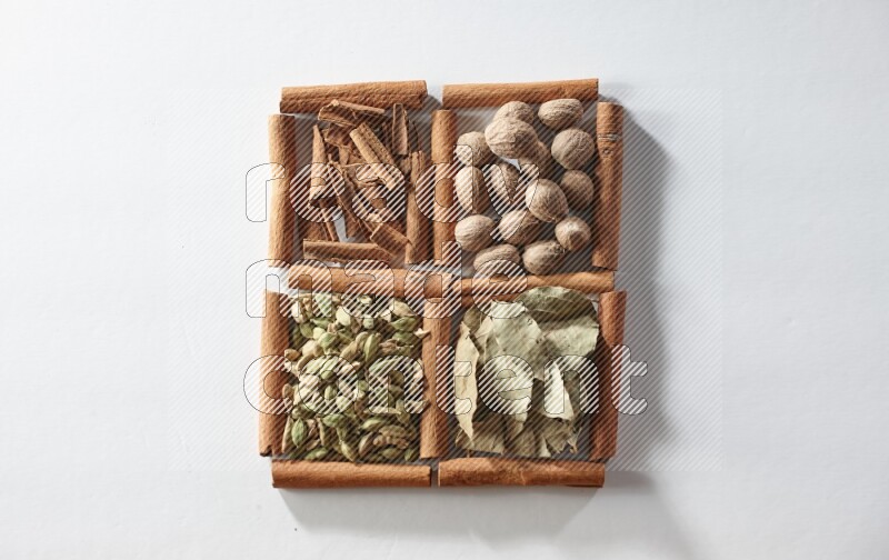 4 مربعات من أعواد القرفة ممتلئة بجوزة الطيب وأوراق الغار وحبوب الهيل والقرفة على أرضية بيضاء