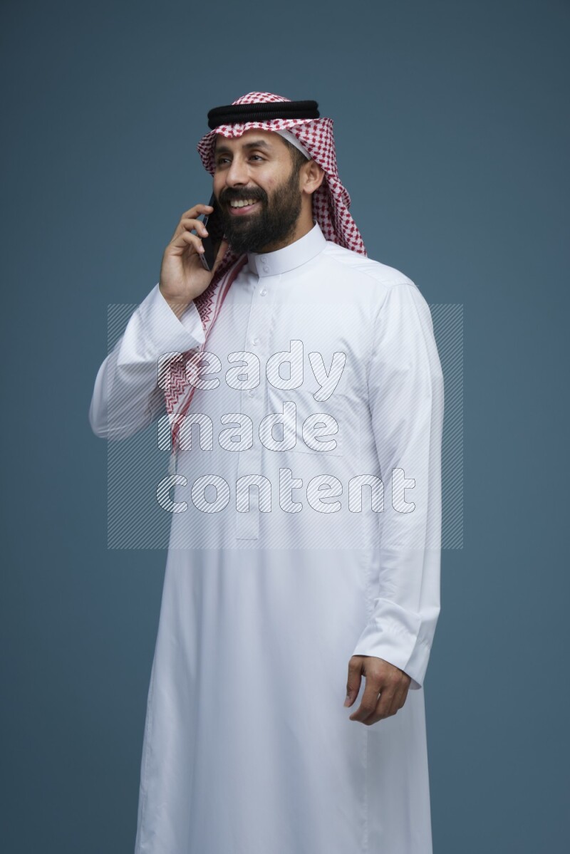 A man having a Call in a blue background wearing Saudi Thob with Shomag
