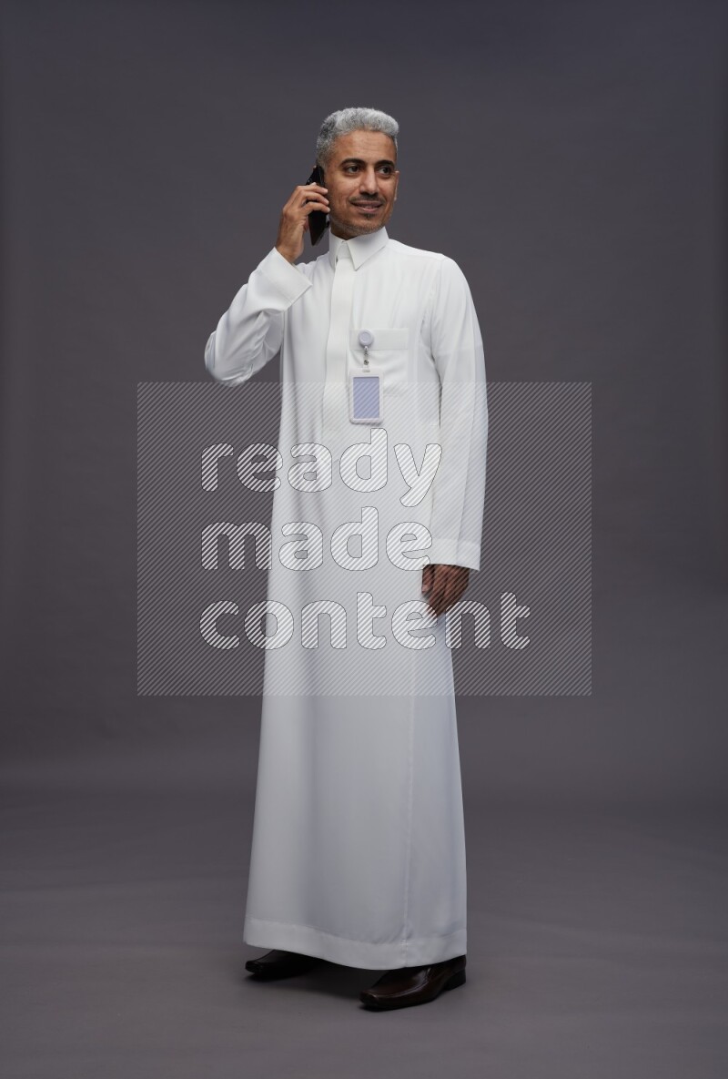 Saudi man wearing thob with pocket employee badge standing talking on phone on gray background