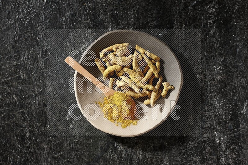 A plate filled with dried turmeric fingers and a wooden spoon full of turmeric powder on a textured black flooring