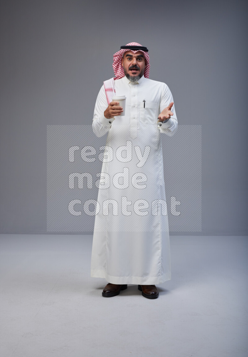 Saudi man Wearing Thob and red Shomag standing holding paper cup on Gray background