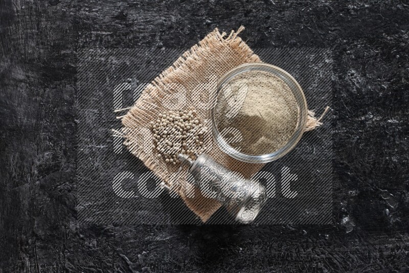 A glass bowl full of white pepper powder with white pepper beads on a burlap piece of fabric and a metal grinder on textured black flooring