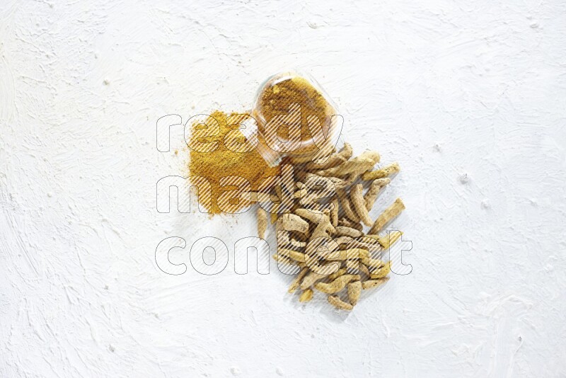 A flipped glass spice jar full of turmeric powder and powder spilled out of it with dried whole fingers on textured white flooring